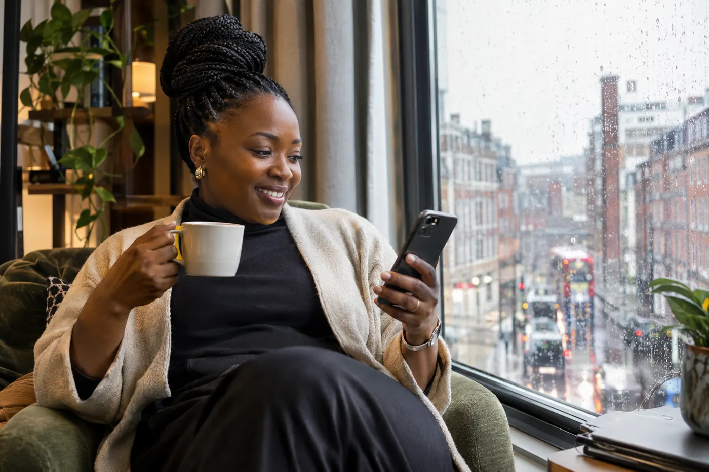 A woman abroad smiling at her phone with a coffee by a rainy city window