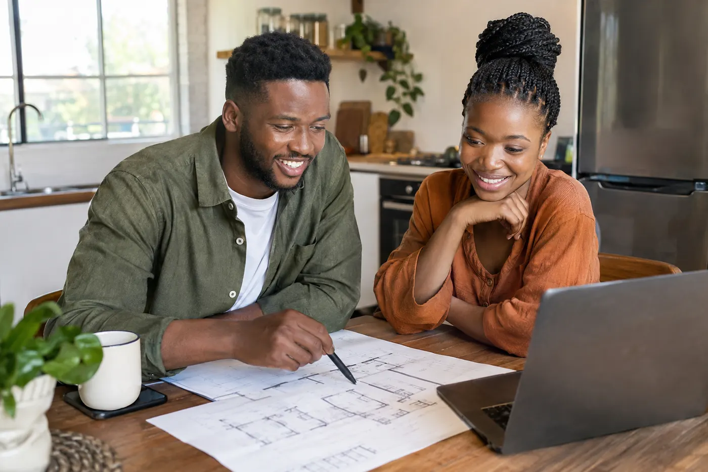 A young couple smiling over building plans and a laptop at their kitchen table